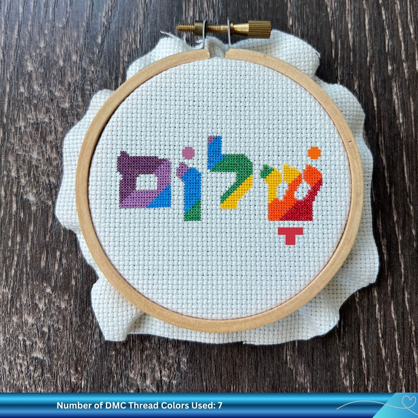 Embroidery hoop with colorful Hebrew word cross-stitched text on a wooden surface, surrounded by embroidery threads and scissors.
