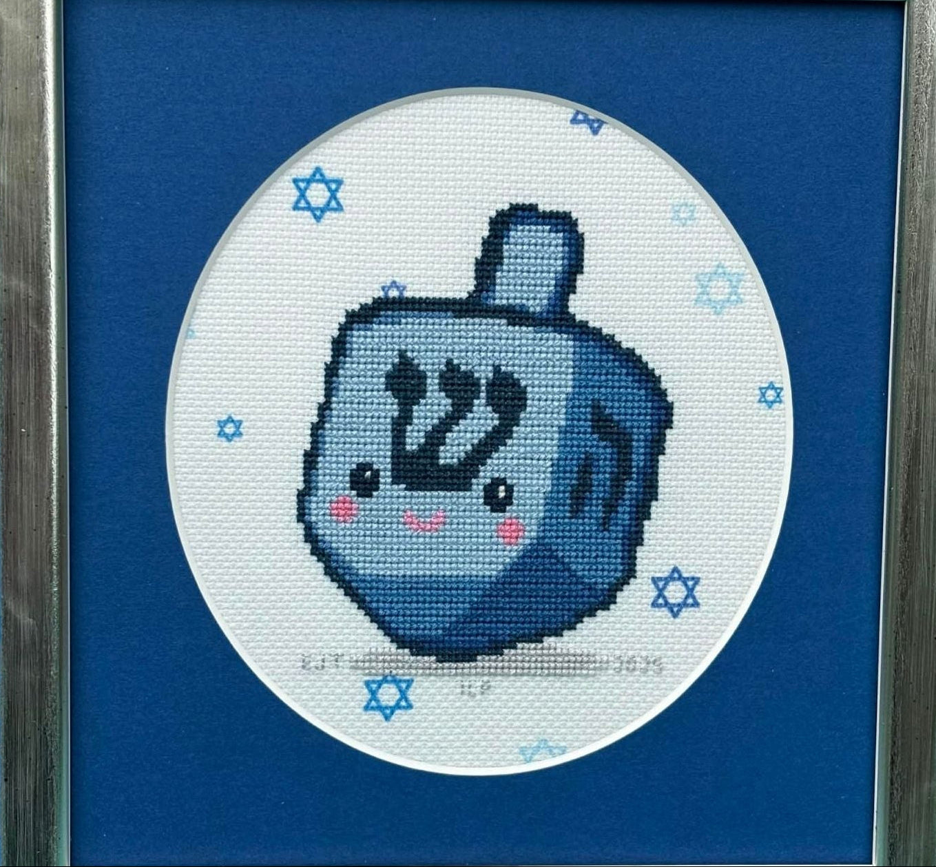 Cross-stitched blue dreidel with a happy face on a white background with blue stars of David.