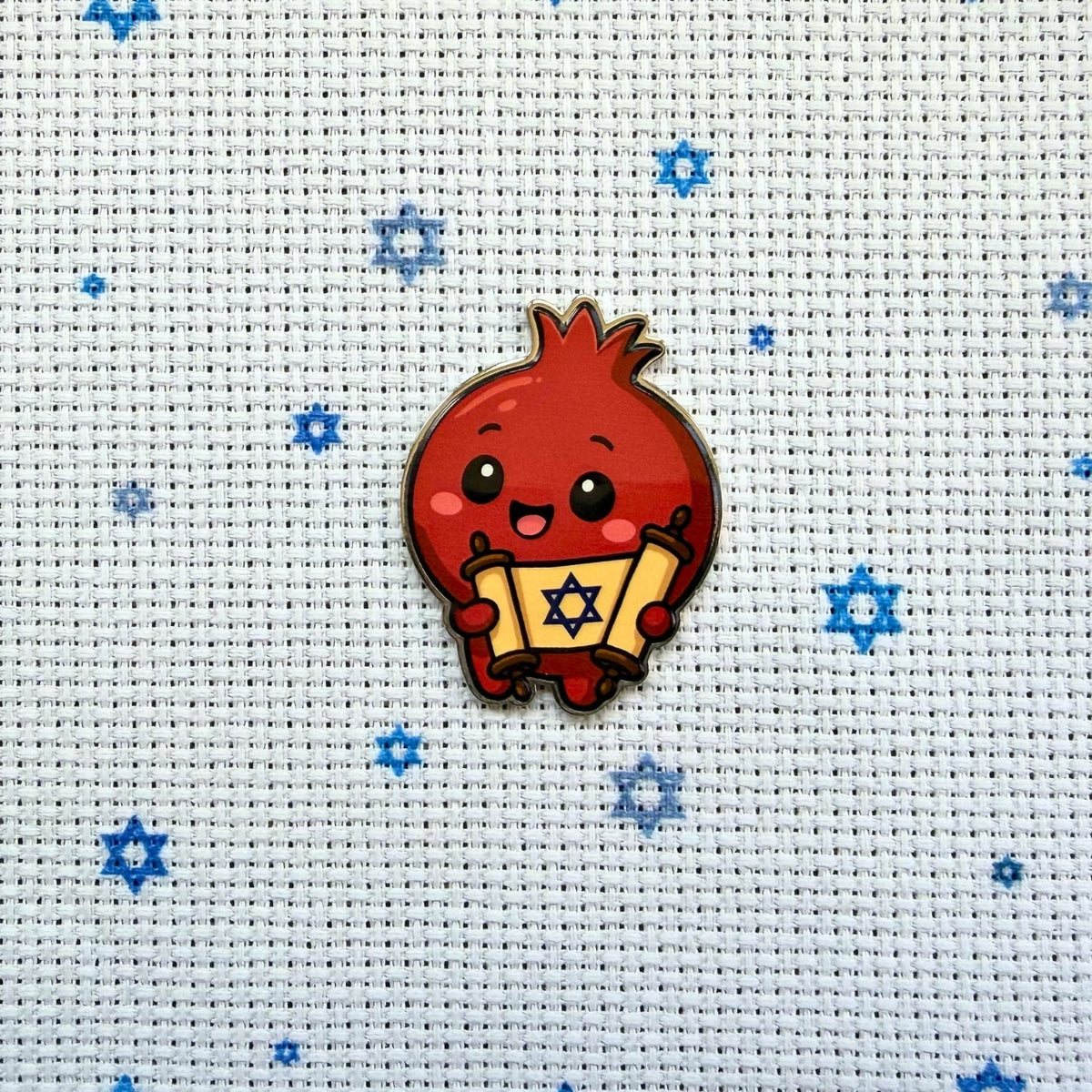 Cute cartoon pomegranate character holding a scroll with a star on a cross-stitched background.