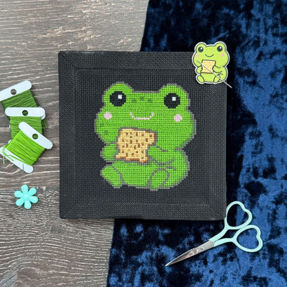 Cross-stitched frog design on a black fabric with green thread, scissors, and embroidery thread on a wooden surface.