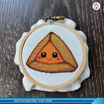 Cross-stitched design of a smiling apricot hamantaschen character inside a wooden embroidery hoop.