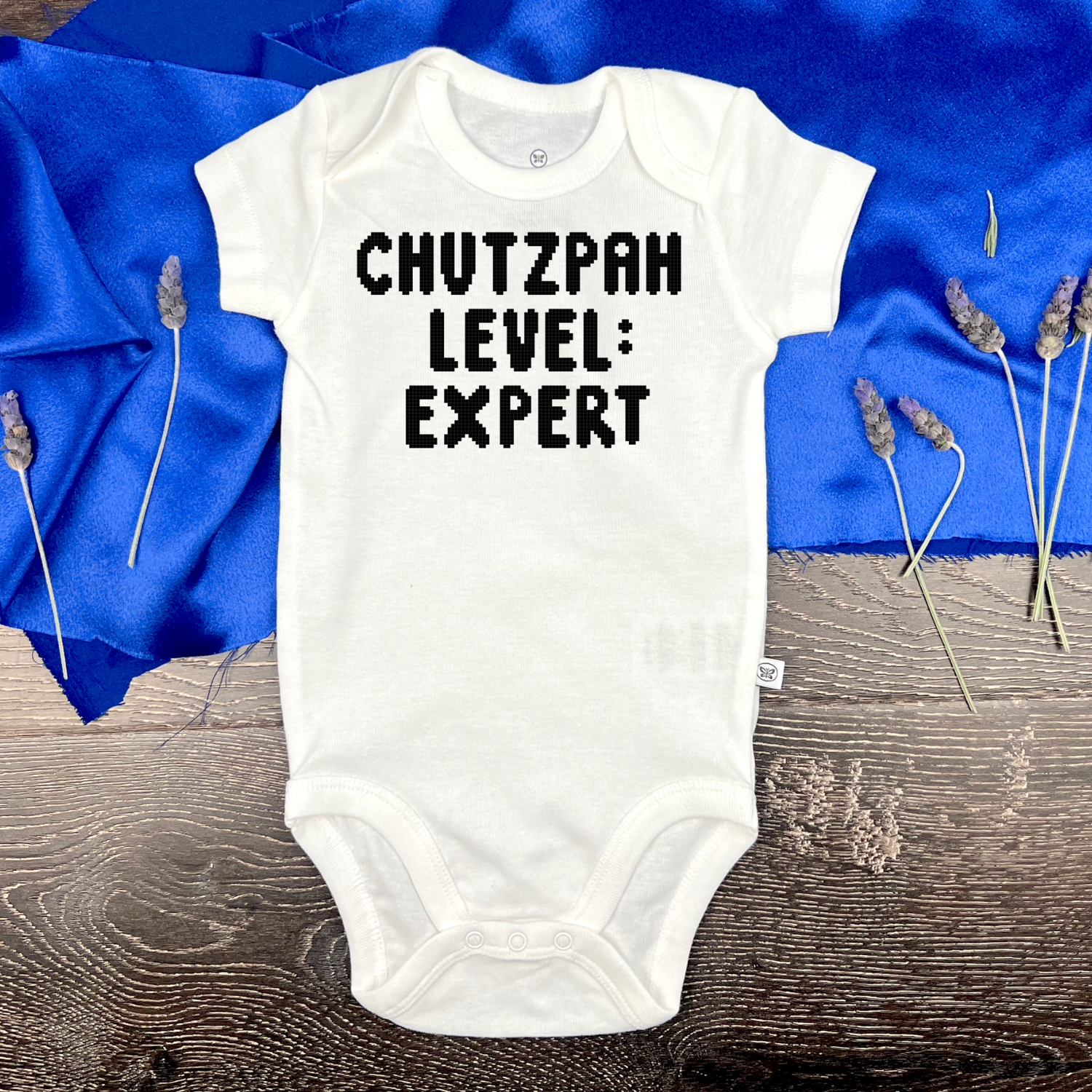White baby onesie with 'CHUTZPAH LEVEL: EXPERT' text on a blue fabric background.