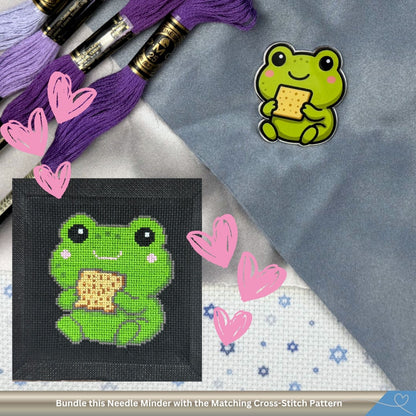 Cross-stitched frog pattern with matching needle minder on a textured surface with embroidery threads and hearts.