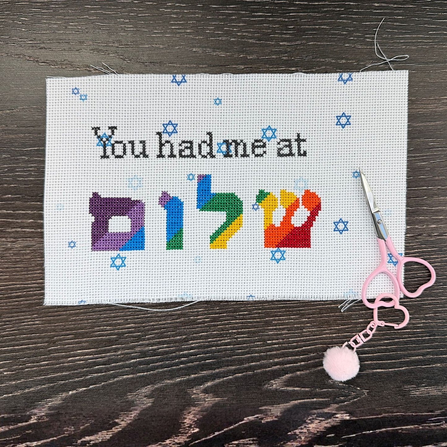 Cross-stitched project with colorful letters and text on a wooden surface with scissors and a pom-pom fob.