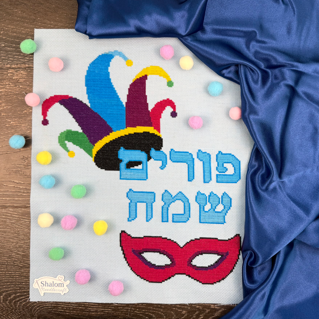 Cross-stitched design for Purim with colorful jester hat, mask, and Hebrew text on a wooden surface.