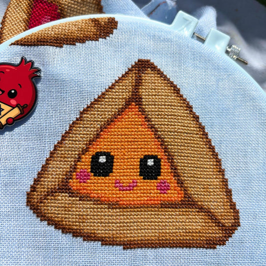 Cross-stitched design of a smiling orange hamantaschen character on a white linen background.