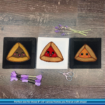 Three embroidered triangular hamantaschen with faces on a wooden surface with scissors and lavender.