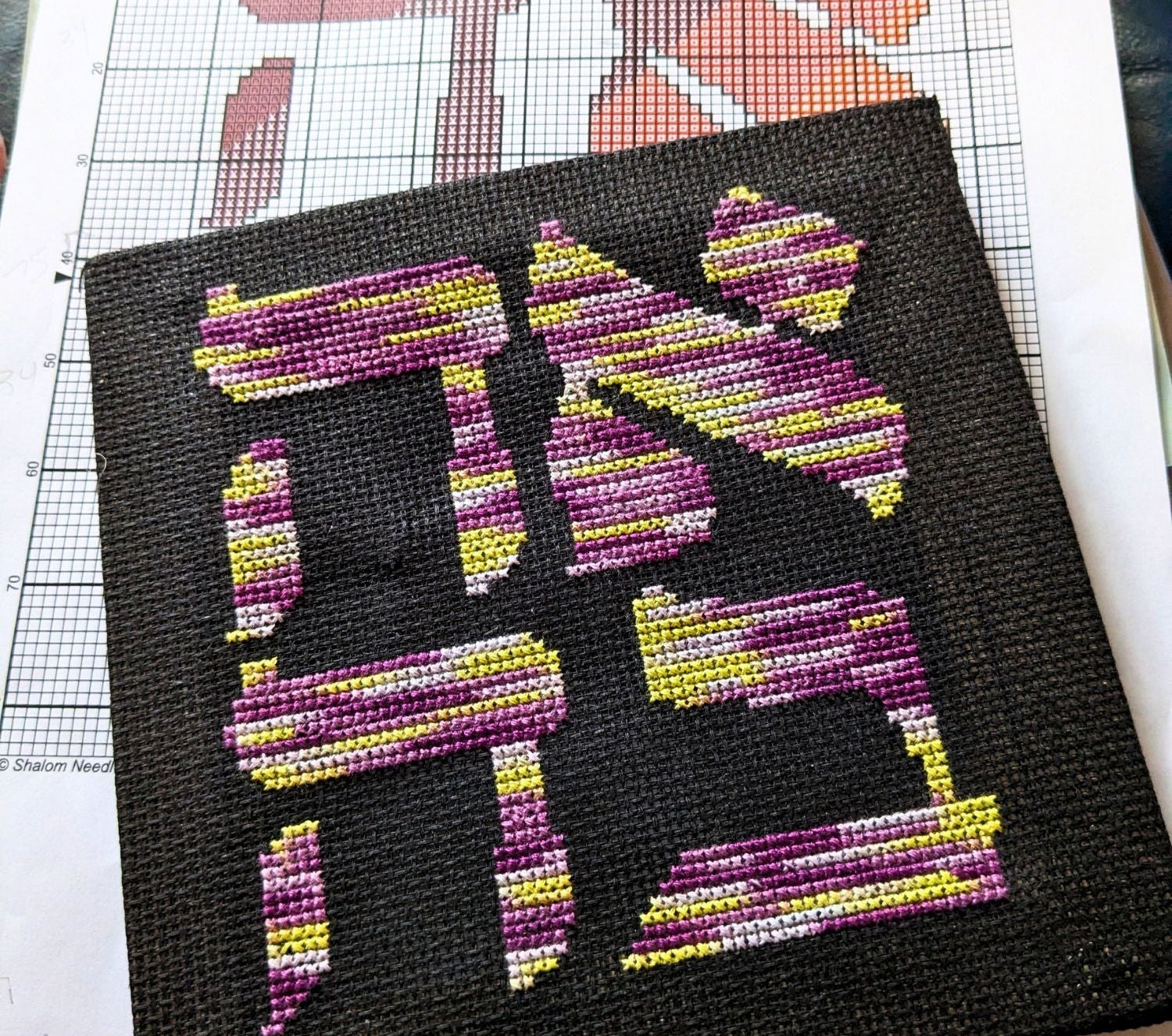 Cross-stitched design of the Hebrew word for Love (Ahava) with colorful letters Hebrew on black fabric, placed on a printed out pattern.