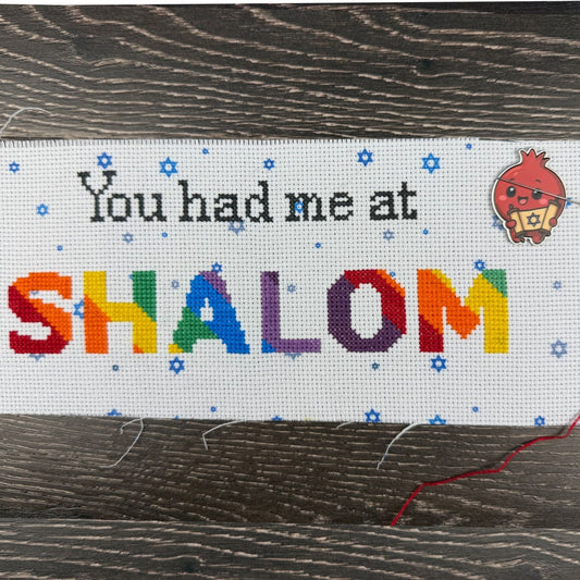 Cross-stitched text 'You had me at SHALOM' with colorful letters on a wooden surface.