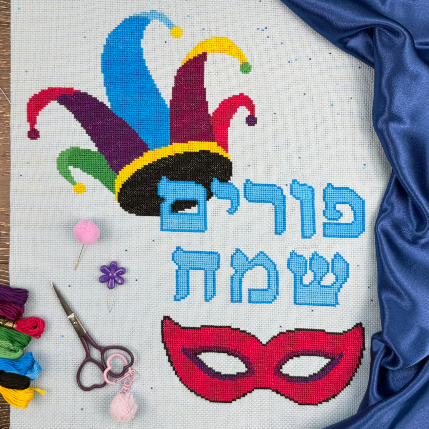 Jewish chag Purim cross-stitched design with colorful jester hat and mask on fabric, surrounded by embroidery tools.