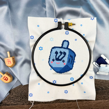 Cross-stitching project with a blue dreidel design on white fabric, surrounded by wooden dreidels and a blue fabric background.