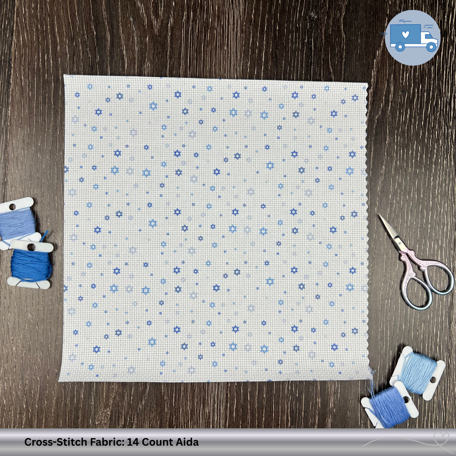Cross-stitch fabric with blue Star of David pattern on a wooden surface, surrounded by sewing tools.