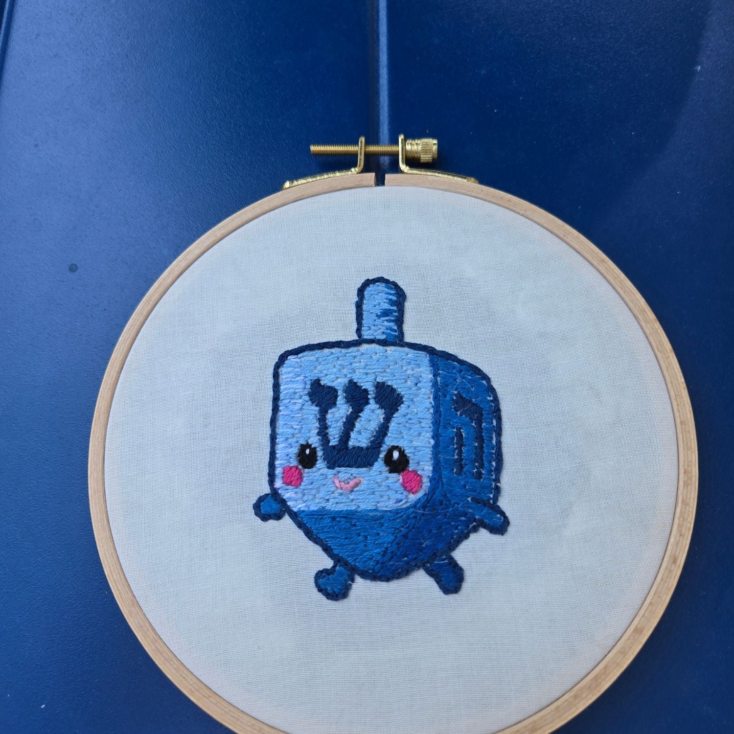 Embroidery of a Hanukkah dreidel with chibi face in a wooden hoop on a blue background