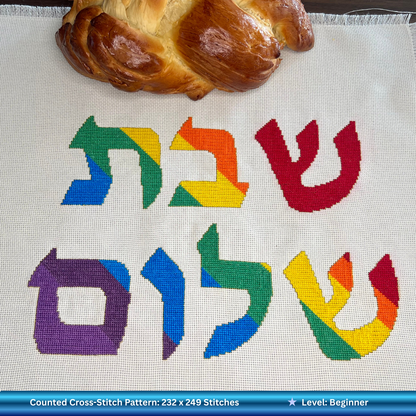 Cross-stitched rainbow Hebrew letters on fabric with a loaf of Challah bread