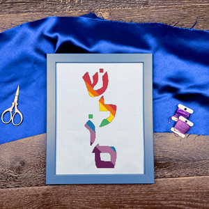 Framed cross-stitched artwork with colorful letters on a blue fabric background