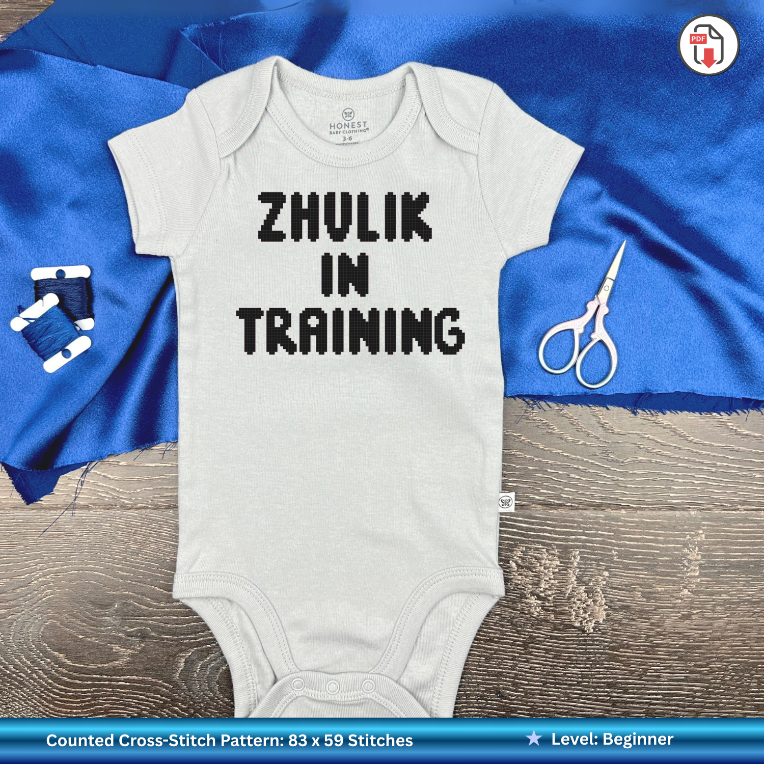 White baby onesie with 'ZHULIK IN TRAINING' text on a blue fabric background.