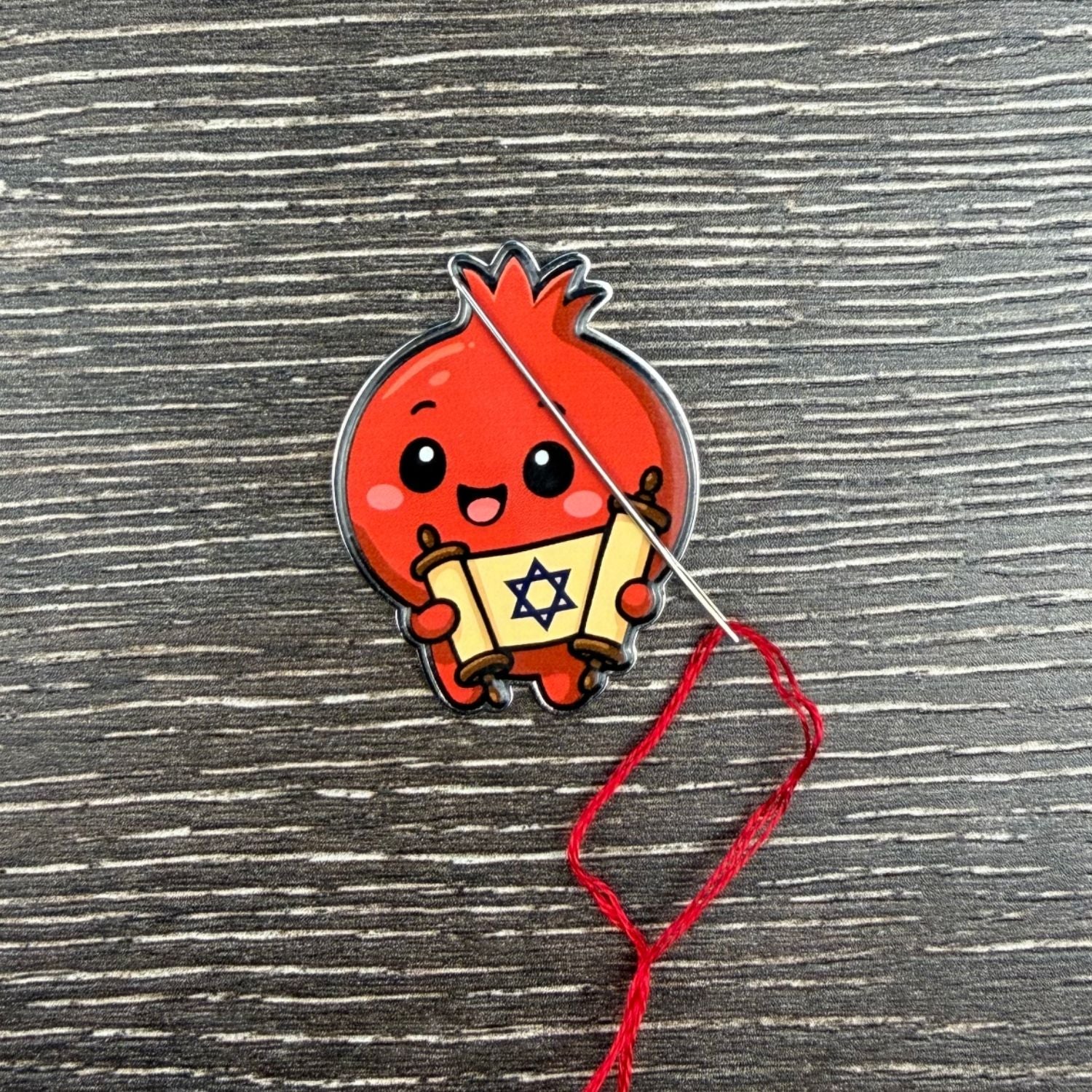 Red cartoon-like pomegranate holding a Jewish Torah scroll with a star design on a cross-stitch background.