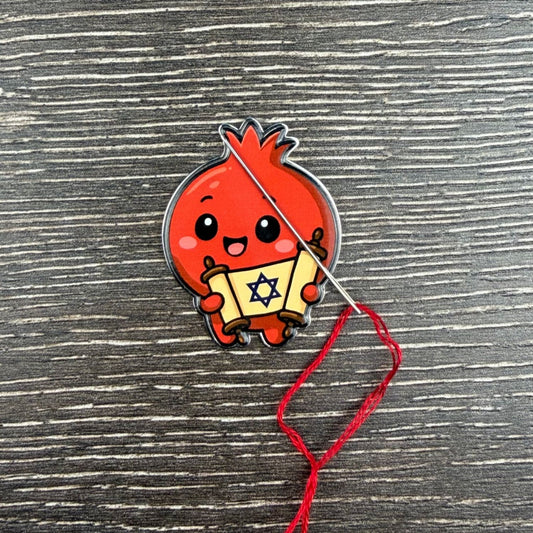 Red cartoon-like pomegranate holding a Jewish Torah scroll with a star design on a cross-stitch background.