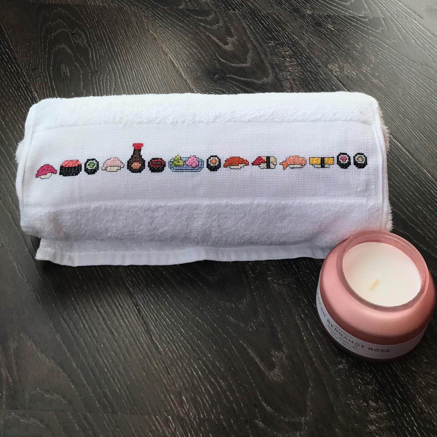 White towel with sushi-themed cross-stitch design on a wooden surface with a pink candle.