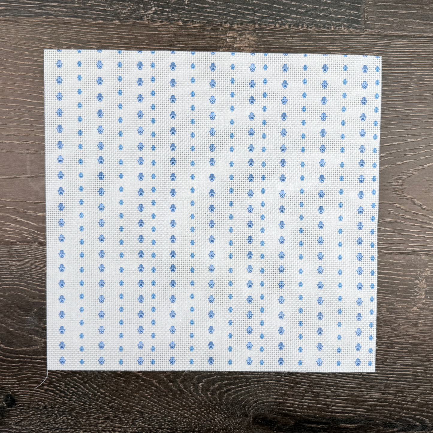 Piece of cotton Aida with blue Hamsa symbols printed lying against a wooden background