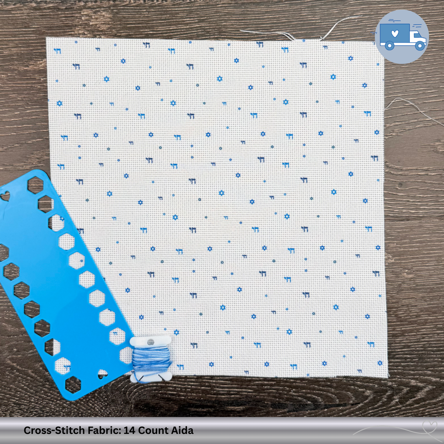 Cross-stitch fabric with blue Chai and Magen David symbols, a blue plastic thread organizer, and a spool of thread on a wooden surface.