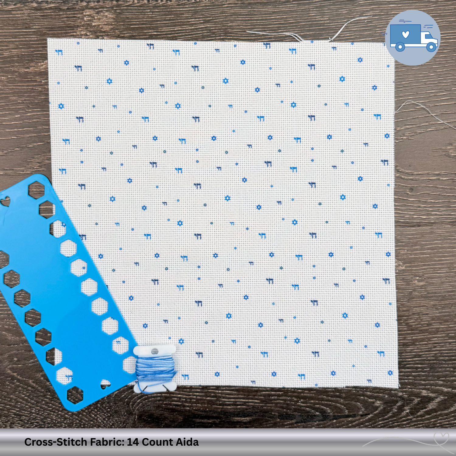 Cross-stitch fabric with blue Chai and Magen David symbols, a blue plastic thread organizer, and a spool of thread on a wooden surface.