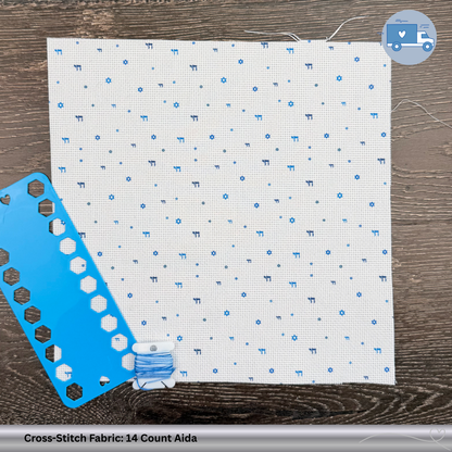 Cross-stitch fabric with blue Chai and Magen David symbols, a blue plastic thread organizer, and a spool of thread on a wooden surface.