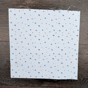 Piece of speciality cotton Aida fabric with blue stars of david and chai symbols printed lying against a wooden background