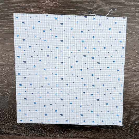 Piece of speciality cotton Aida fabric with blue stars of david and chai symbols printed lying against a wooden background