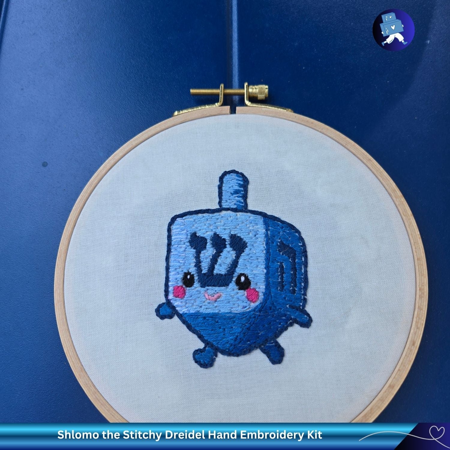 Embroidery hoop with a blue embroidered dreidel on a blue background, featuring 'Shlomo the Stitchy Dreidel Hand Embroidery Kit'.