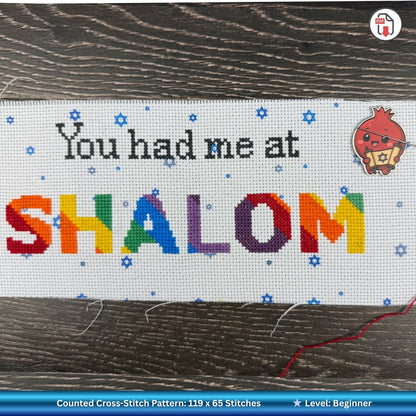Cross-stitched Sampler with 'You had me at SHALOM' text and pomegranate needle holder on wooden background