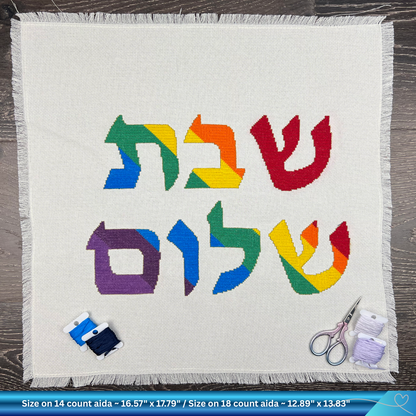 Colorful Hebrew text saying Shabbat Shalom on a white fabric with embroidery tools on a wooden surface
