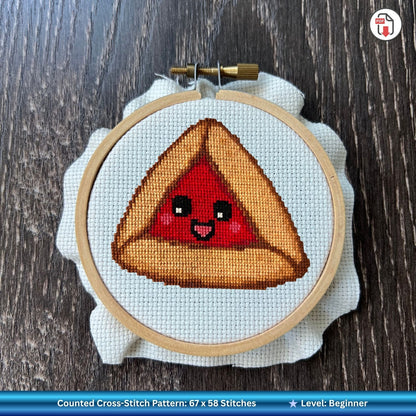 Cross-stitched design of a smiling raspberry hamantaschen character inside embroidery hoop.