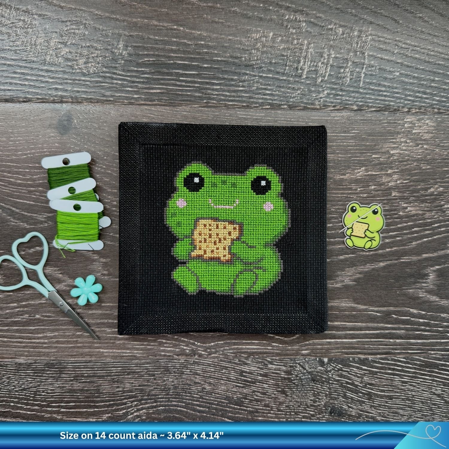 Cross-stitched frog design on a black fabric Aida square with green thread and scissors on a wooden surface.