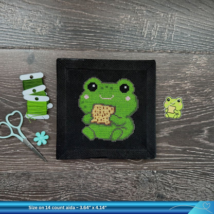 Cross-stitched frog design on a black fabric Aida square with green thread and scissors on a wooden surface.