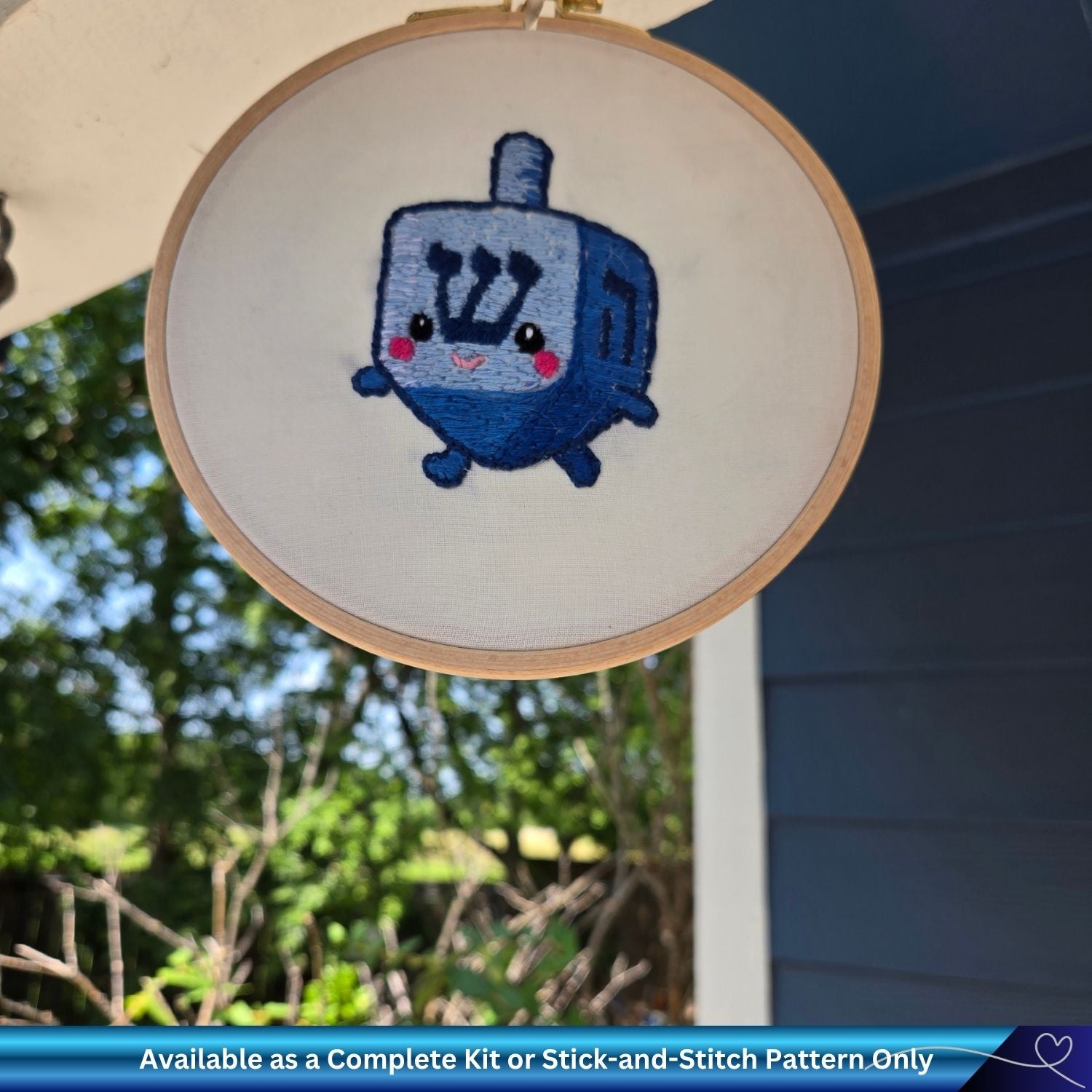 Embroidery hoop with a blue embroidered dreidel on a white background, outdoors.