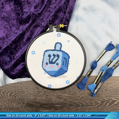 Embroidery hoop with a blue dreidel design on white fabric, surrounded by embroidery threads and cloths.