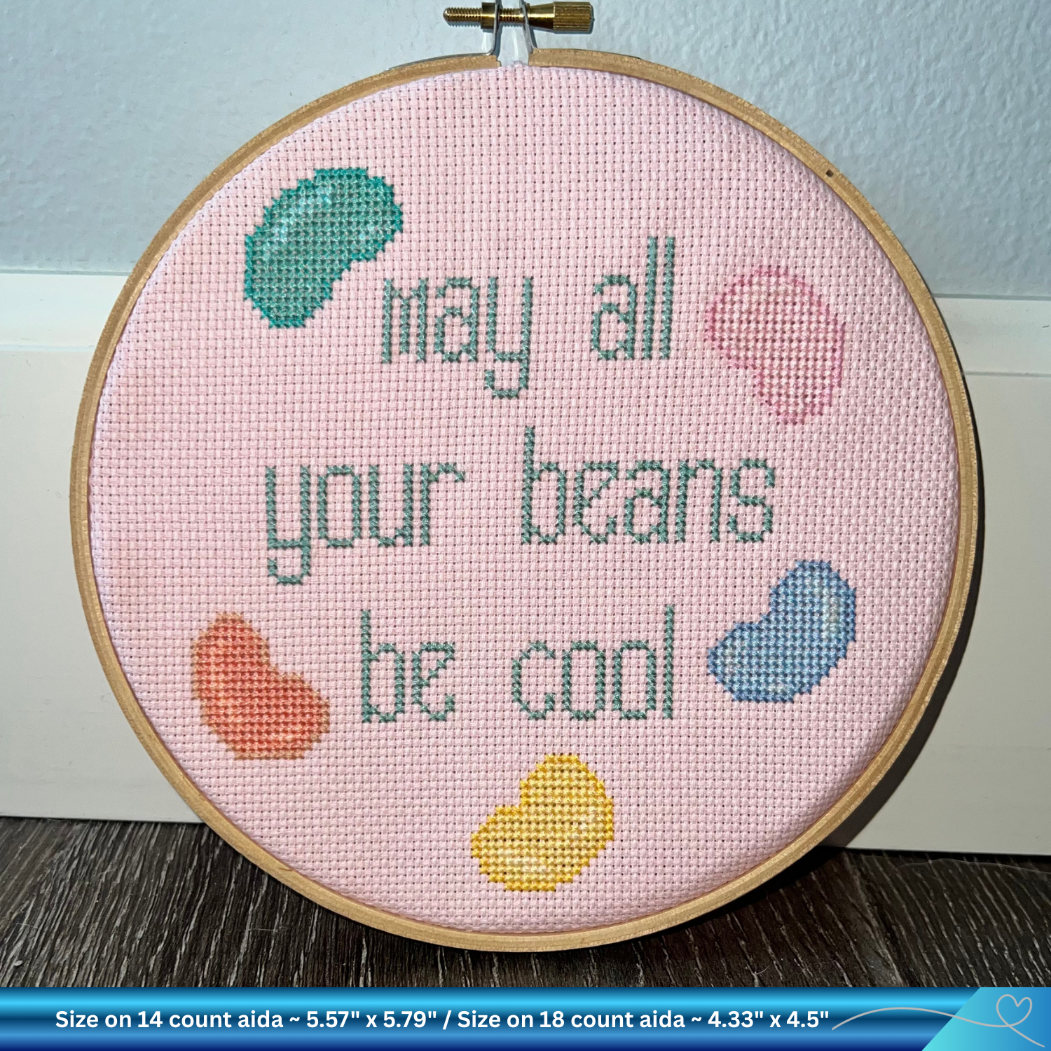 Cross-stitched hoop with 'May all your beans be cool' text and colorful beans on a pink background.