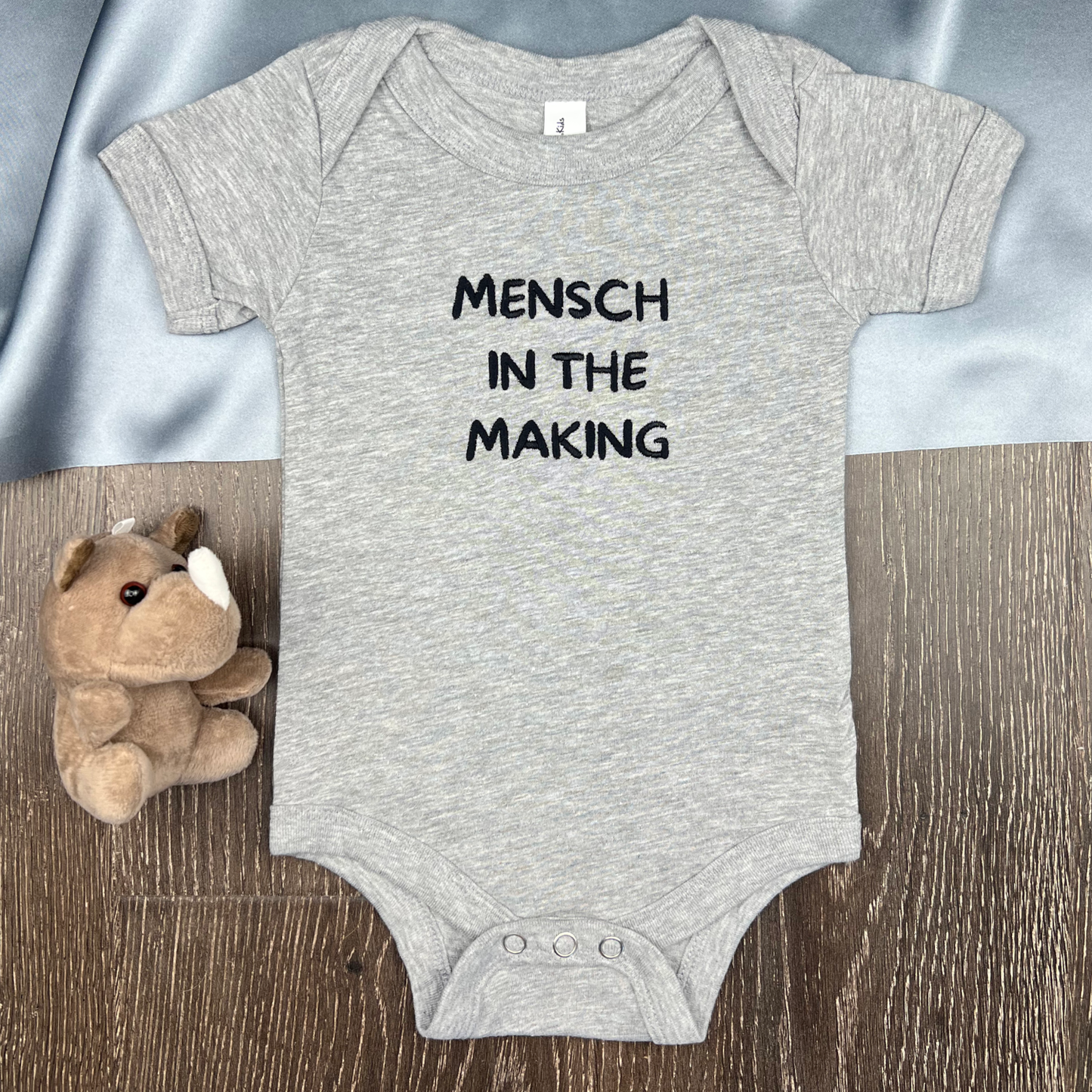 Gray baby onesie with 'Mensch in the Making' text embroidered on the front against a wooden surface with a plush toy.