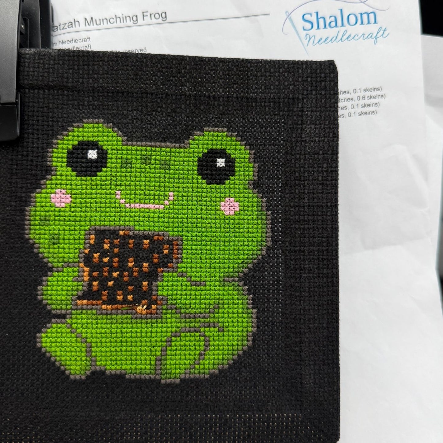 Cross-stitched green frog on black fabric with 'Shalom Needlecraft' branding in the background.