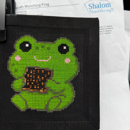 Cross-stitched green frog on black fabric with 'Shalom Needlecraft' branding in the background.