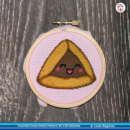 Cross-stitched hamantaschen with a face in an embroidery hoop on a wooden surface