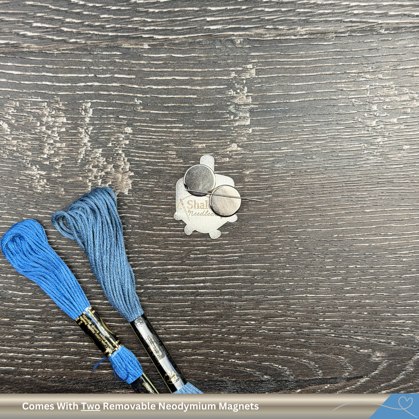 Two embroidery floss spools with a magnetic dreidel needle minder on a wooden surface, featuring the brand 'Shalom Needlecraft'.