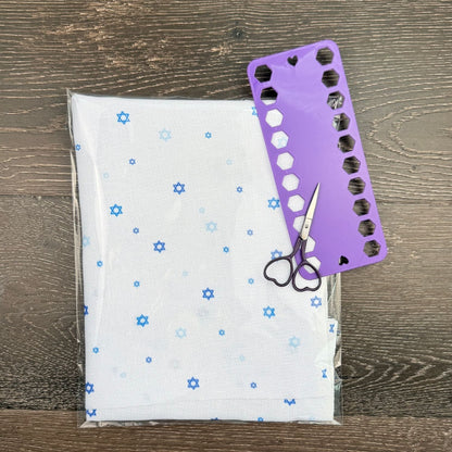 Scissors on a piece of fabric with star of david patterns and a purple floss organizer on a wooden surface