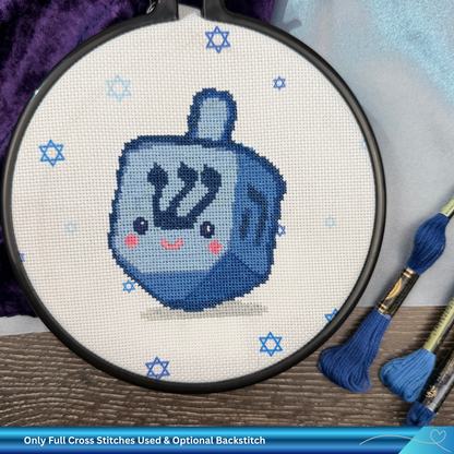 Cross-stitched blue dreidel with pink cheeks in an embroidery hoop, surrounded by blue stars of David.