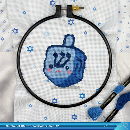 Cross-stitched blue dreidel in an embroidery hoop with DMC thread on a white fabric background.