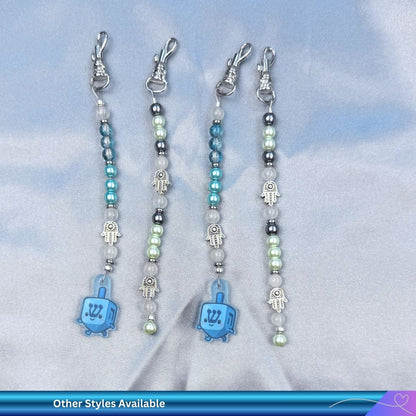 Four beaded keychains with blue dreidel and hamsa charms on a light blue background.