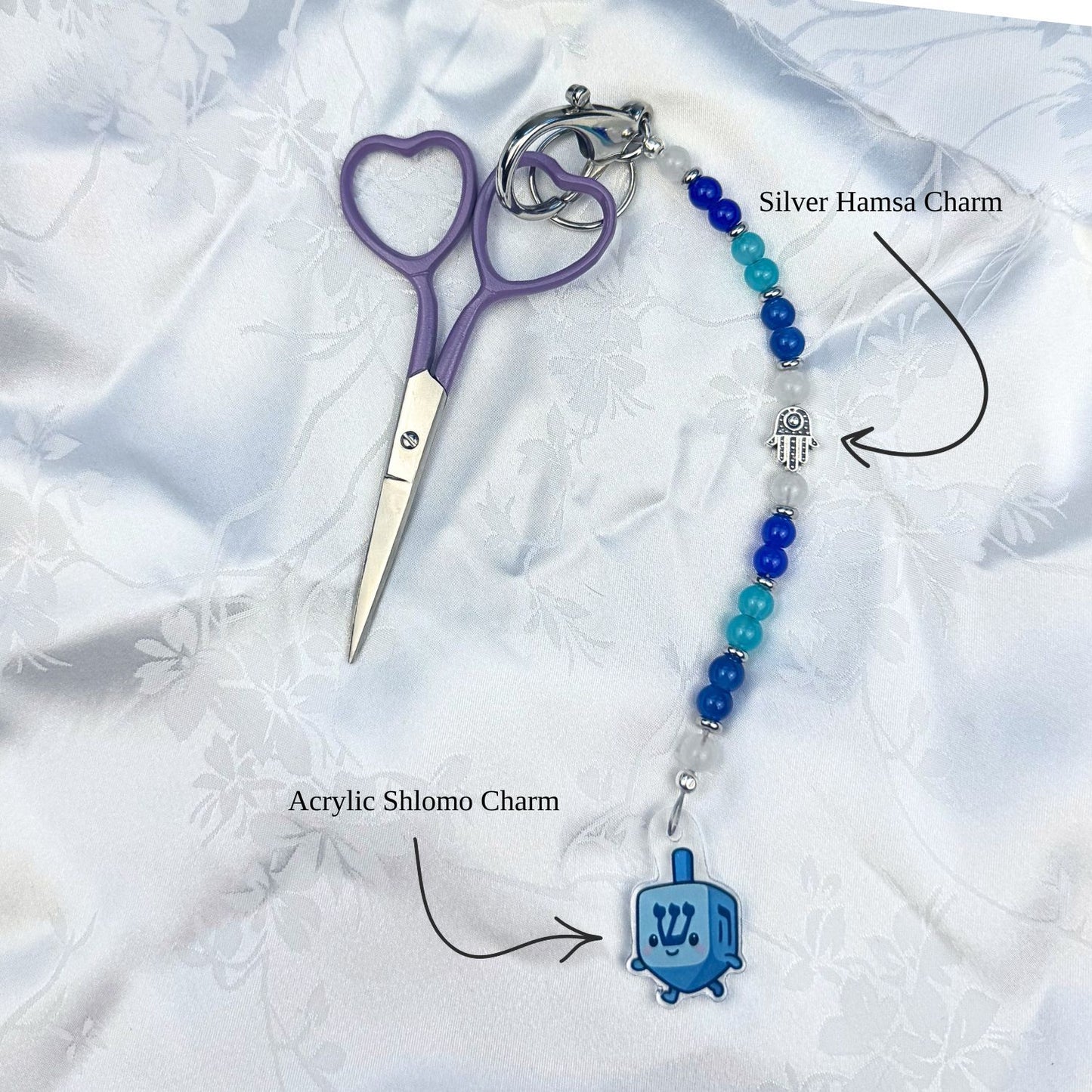 Keychain with heart-shaped scissors, silver hamsa charm, and blue acrylic Shlomo dreidel Hanukkah charm on a white fabric background.