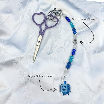 Keychain with heart-shaped scissors, silver hamsa charm, and blue acrylic Shlomo dreidel Hanukkah charm on a white fabric background.