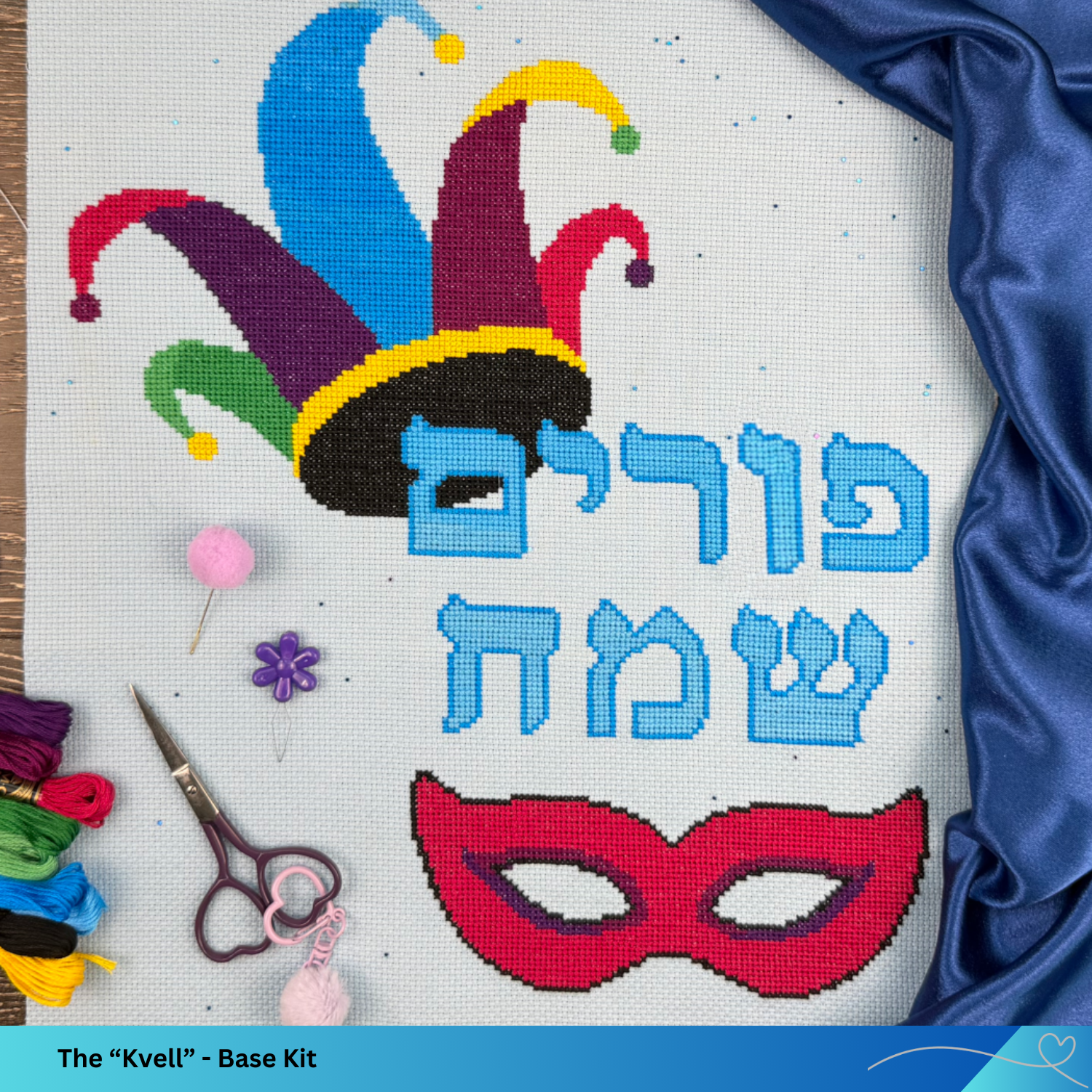 Cross-stitch pattern with colorful jester hat, mask, and Hebrew text on fabric with sewing tools.